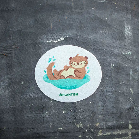 A round kitchen sponge with an illustration of a sleeping otter in a splash of water, surrounded by the text 'PLANTISH'.