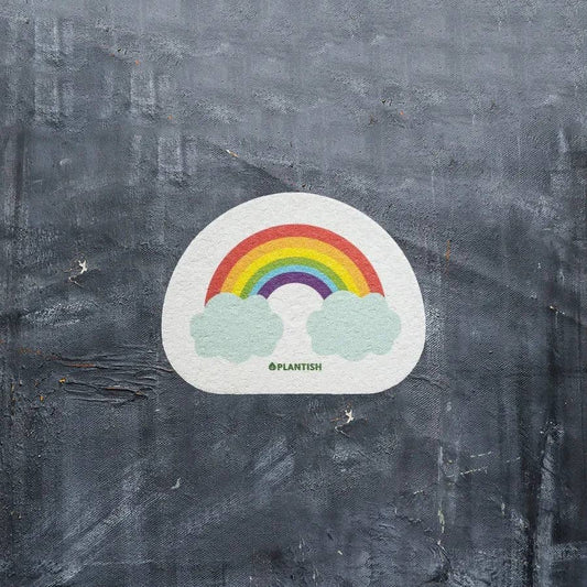 A white sponge with a printed rainbow and cloud design, with the brand 'Plantish' written on it, placed on a grey textured background.